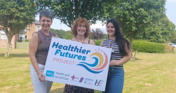 Photograph of three women standing outdoors on grass holding a "Healthier Futures Project" sign with logos for Health and Social Care and HSE. Background includes large green trees and a clear blue sky