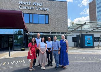 Photograph of six people standing outside North West Cancer Centre, posing in front of building entrance with clear blue sky above. Group includes three men and three women dressed in casual and semi-formal attire, with building signage and modern architecture visible in background.