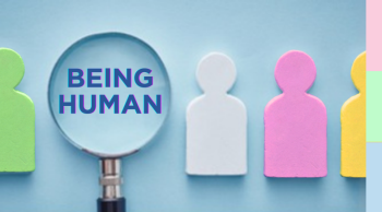 Photograph showing a magnifying glass focusing on a blue figure with text "BEING HUMAN" among a row of colourful human-shaped cutouts in green, white, pink, and yellow. The image symbolises examination or emphasis on human qualities or identity within a diverse group.