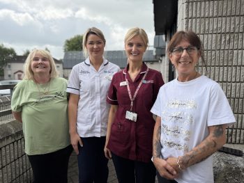 Photograph of four women (Patient Paula, Clare McClintock, Physiotherapist, Maura Ferris, Respiratory Nurse and Patient Leigh) standing outdoors on a balcony with a cloudy sky background. Two women wear medical uniforms in white and maroon, while the other two wear casual clothing, with one displaying tattoos on her arms.