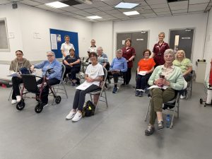 Photograph of a group of individuals seated in a spacious room, accompanied by healthcare workers standing behind them. The setting is a community centre, with participants holding papers and some using mobility aids.