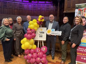 Large group of people standing with pink and yellow balloon arch and sign that displays the words "Adoption Week 2026"