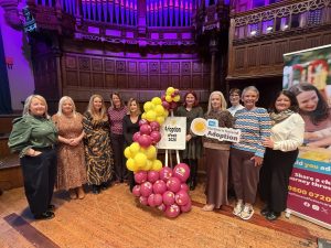 Large group of people standing with pink and yellow balloon arch and sign that displays the words "Adoption Week 2026"